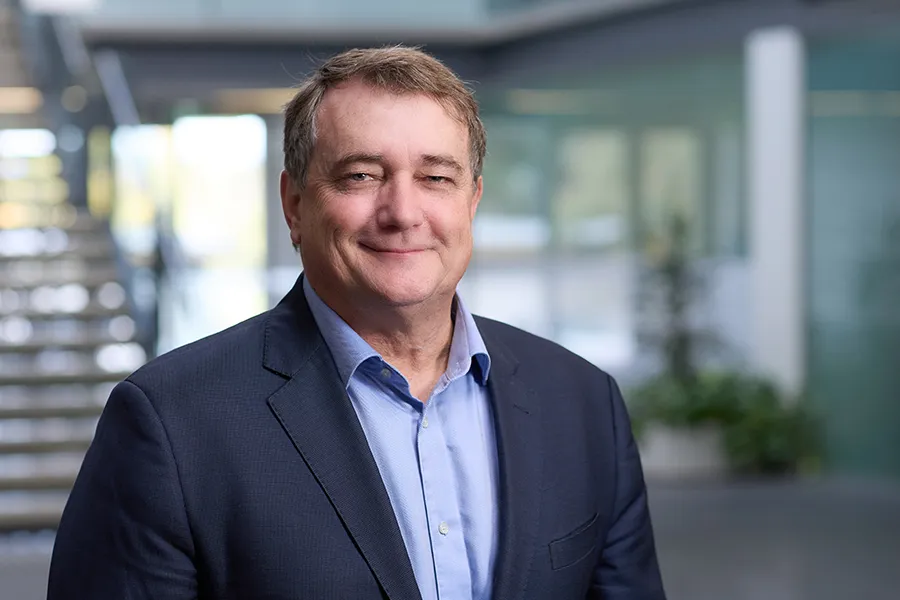 Craig Fitzgerald, Group CEO of Aspen Medical, wearing a navy suit and light blue shirt, smiling in a professional office setting with a modern staircase and glass windows in the background.