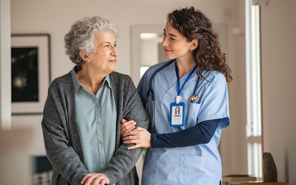 Photo of an elderly women being helped along by a smiling nurse. The nurse has her arm through the womans, to support her.