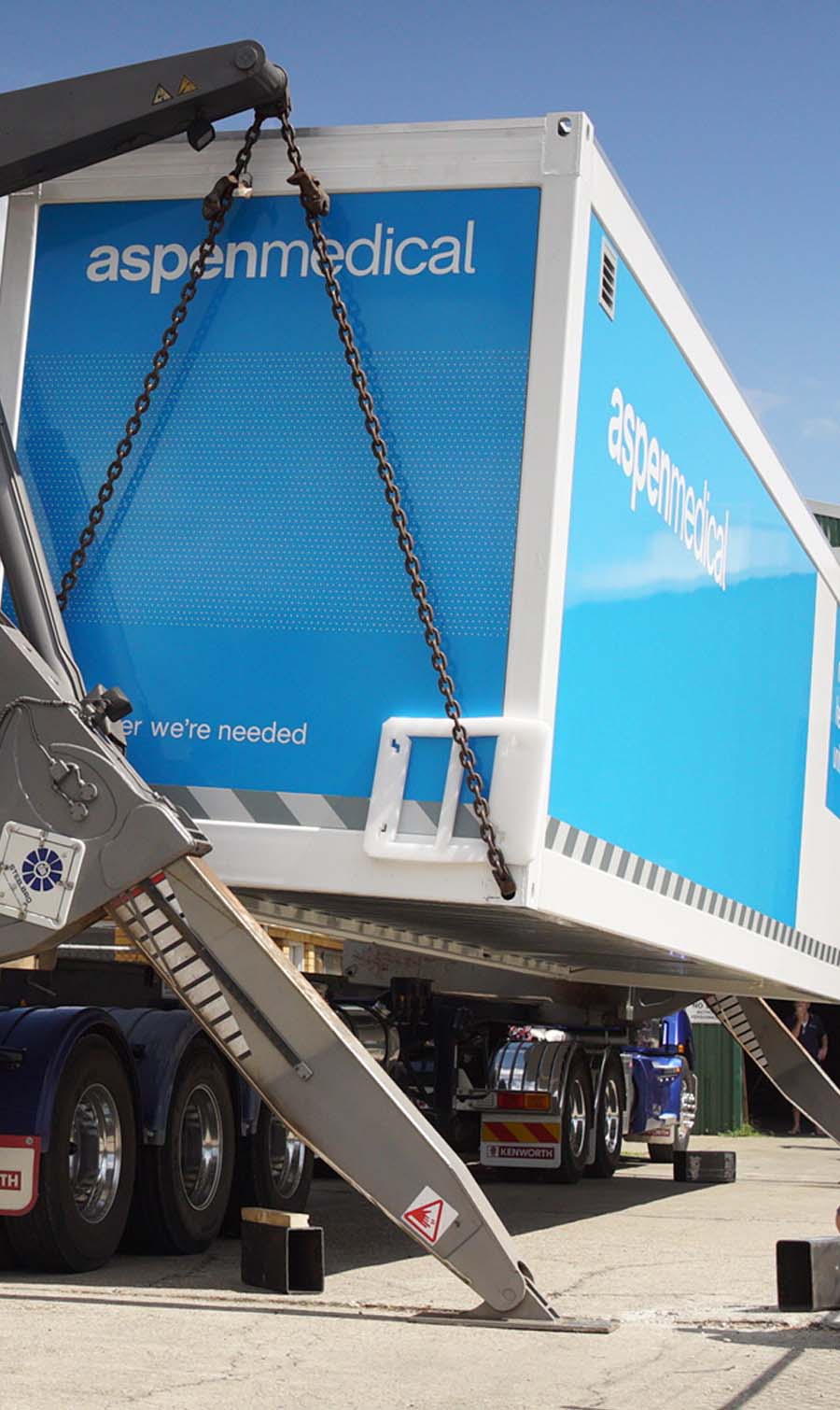 Photo shows an Aspen Medical mobile health unit being hoisted up onto a truck for transportation.