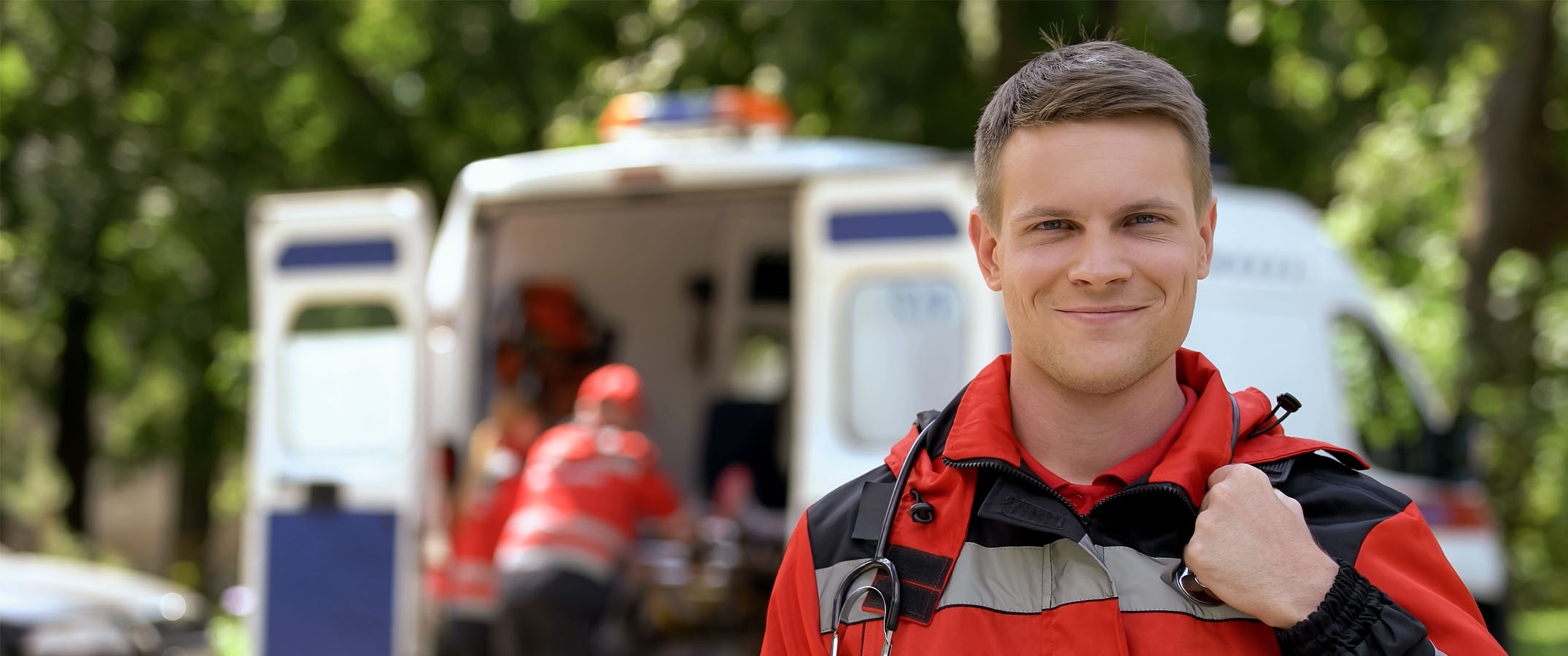 Photo of a male paramedic holding his bag on his shoulder, with an ambulance being loaded in the distance.