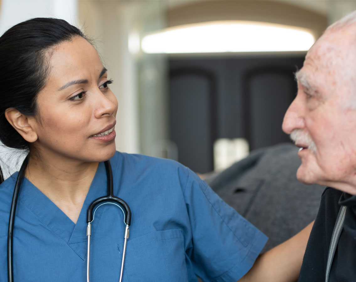 Image shows a nurse to the left looking with care at an elderly gentleman who is talking to her.