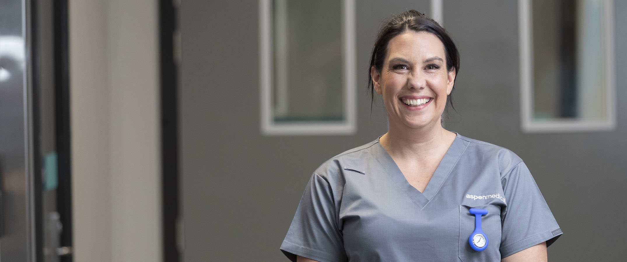 Photo of an Aspen Medical female clinician with dark hair in a ponytail wearing grey scrubs and smiling at the camera.