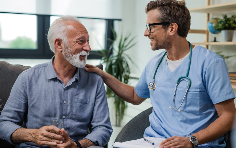 Image of an elderly man with a beard and blue shirt, sitting down next to and looking at a male clinician wearing light blue scrubs, with his hand on the mans shoulder.