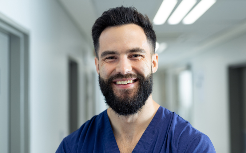 Photo shows a young bearded man with dark hair, wearing a blue top.