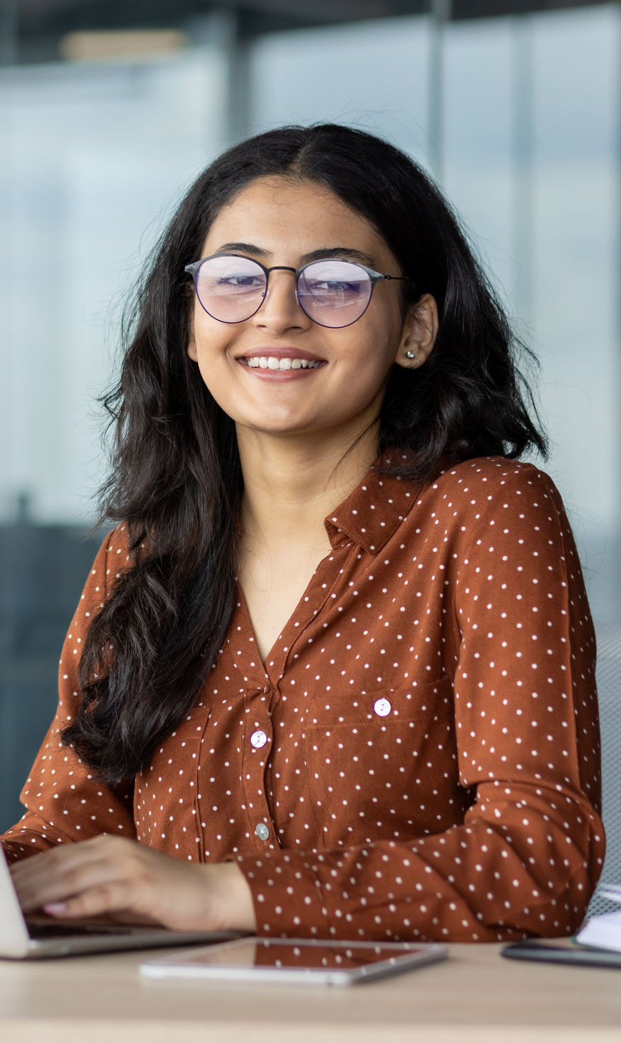 Photo of a professional young woman with long dark hair and glasses, sitting at a desk and smiling at the camera.