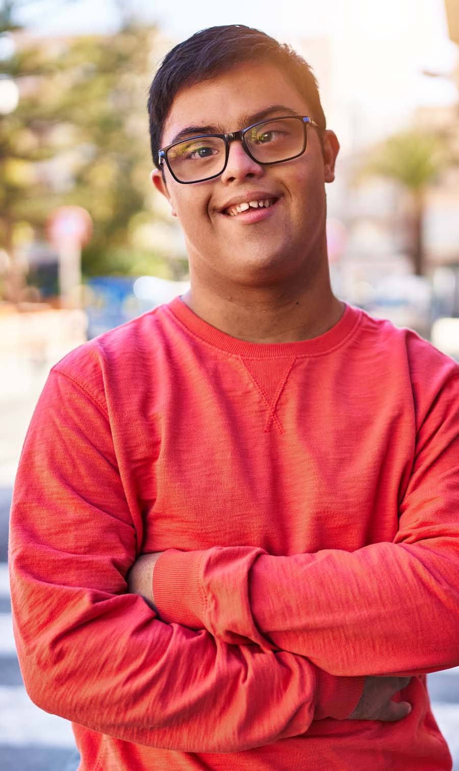 A young man with disability, wearing glasses with arms folded and smiling at the camera.