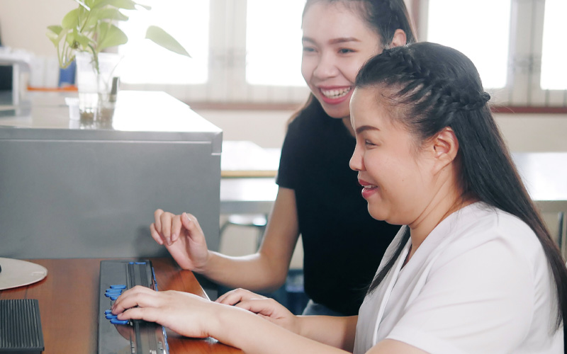 Photo of a blind woman with long dark hair sitting at a computer, using the keyboard with help from another woman.