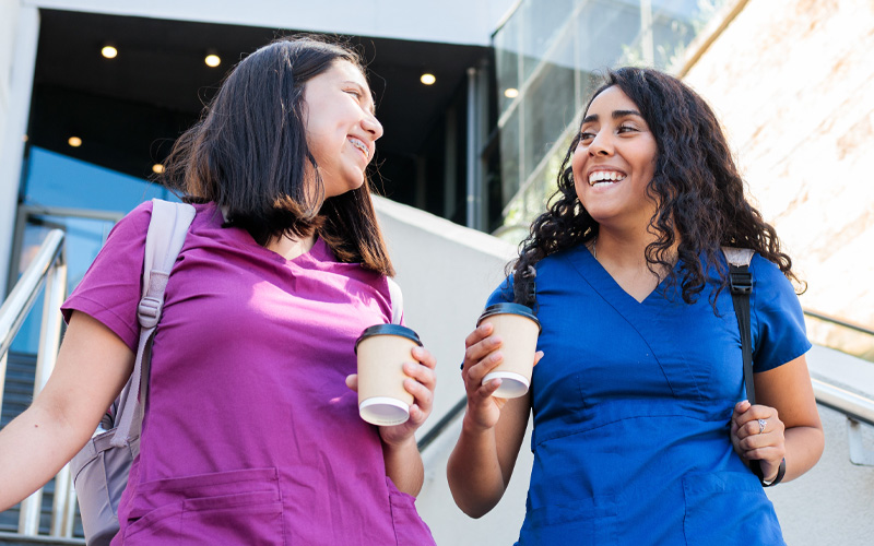 Photo shows two female clinicians walking down the stairs together, smiling and holding coffee cups.