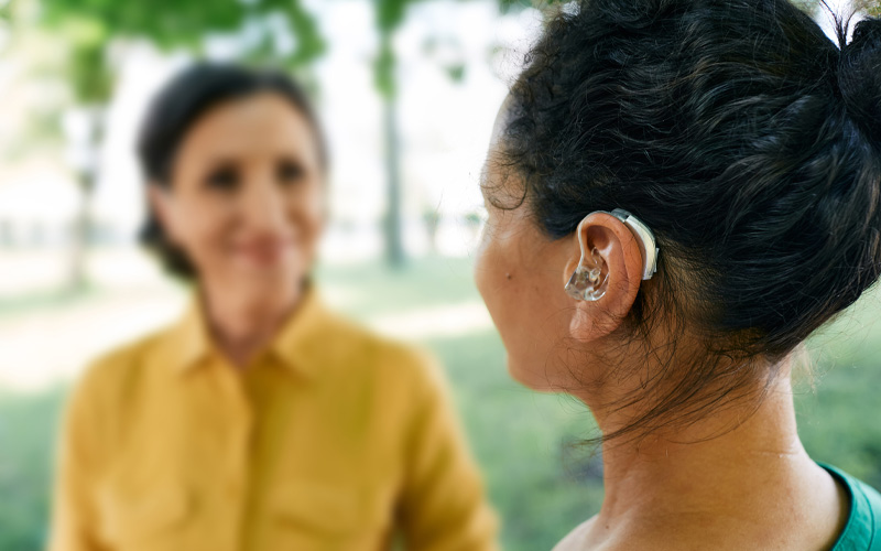 Photo shows a blurry woman in the background, looking at a hearing impaired woman with dark hair who is in profile to us.