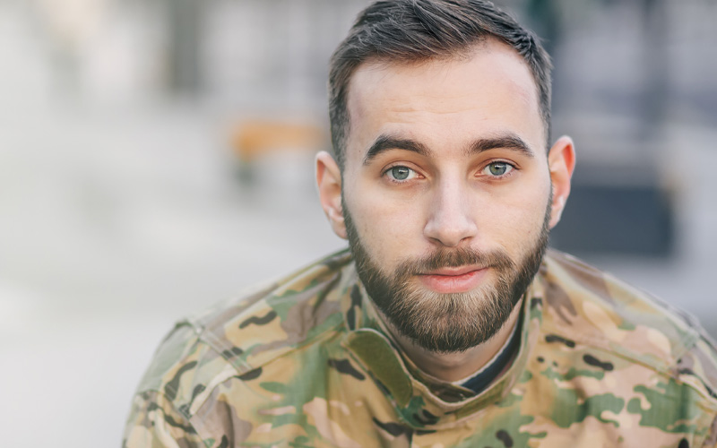 Close up photo of a young man in army fatigues looking at the camera. He has a trimmed beard and dark coloured hair.
