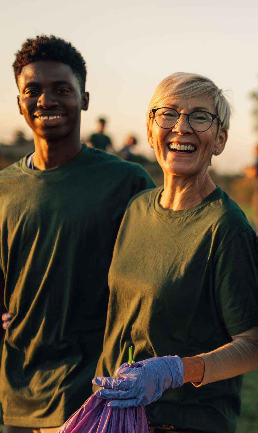 Photo of an older woman and young man standing side by side, smiling and holding a garbage bag. They're outside and the light is golden.