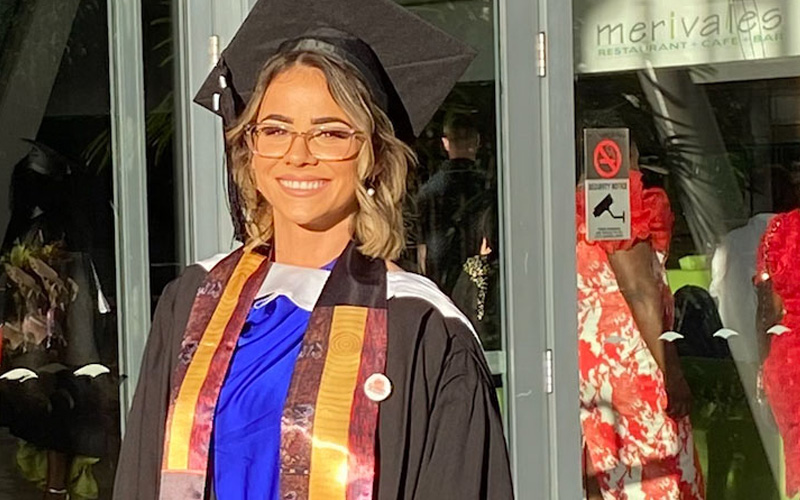 Photo of a young woman with glasses and light hair wearing a graduation gown and cap, smiling at the camera.