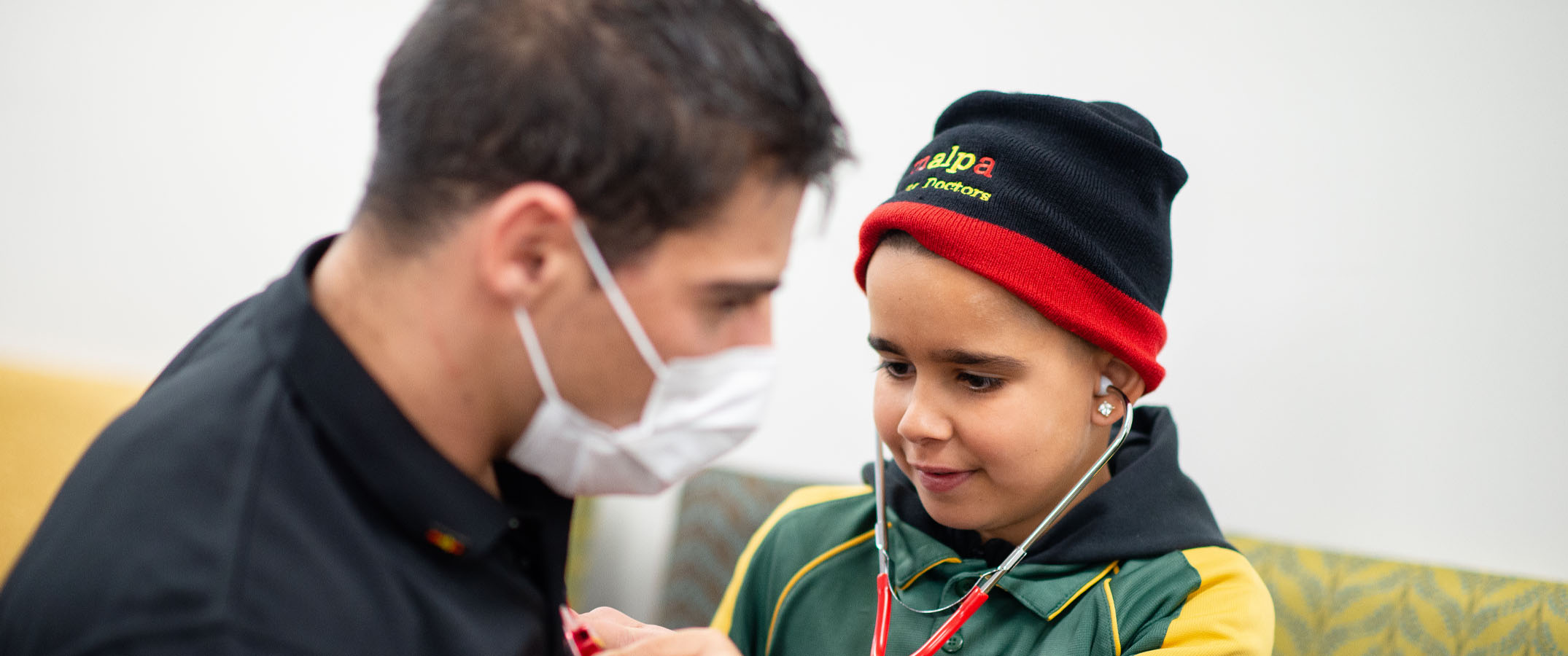 Image of a young Malpa participant, holding a stethoscope to the chest of a man wearing a white mask and black top.