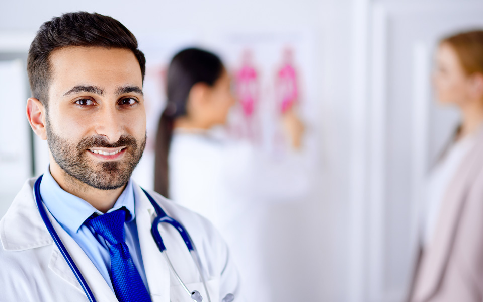 Photo of a male Arabic doctor with a beard with two female clinicians blurred in the background.