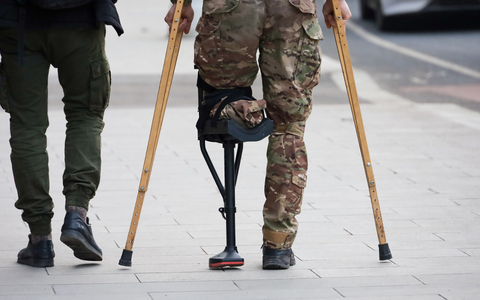 Photo shows two people from the rear, from legs down with the person on the right wearing army fatigues, holding crutches and missing the bottom half of their leg.