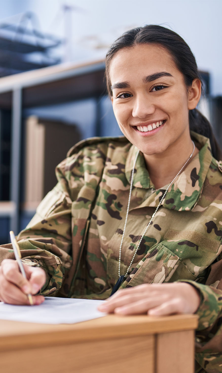 Image of a young, female military personnel, smiling, sitting at a desk and writing.