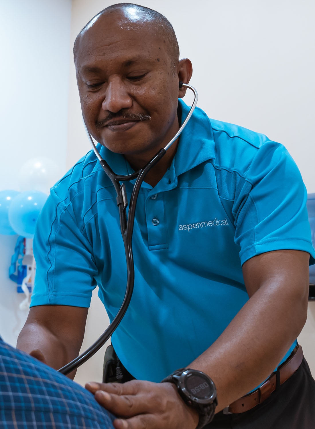 Photo shows an Aspen Medical clinician in PNG tending to a patient, wearing an AM aqua polo.