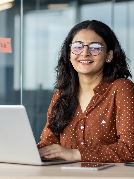 Photo shows a young female office worker with long dark hair, a tan shirt and glasses sitting infront of a laptop.