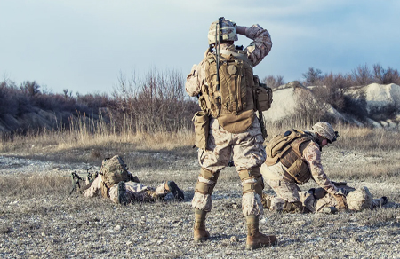Photo shows the back of three soldiers outside, one is tending to the other lying on the group and the other is standing.