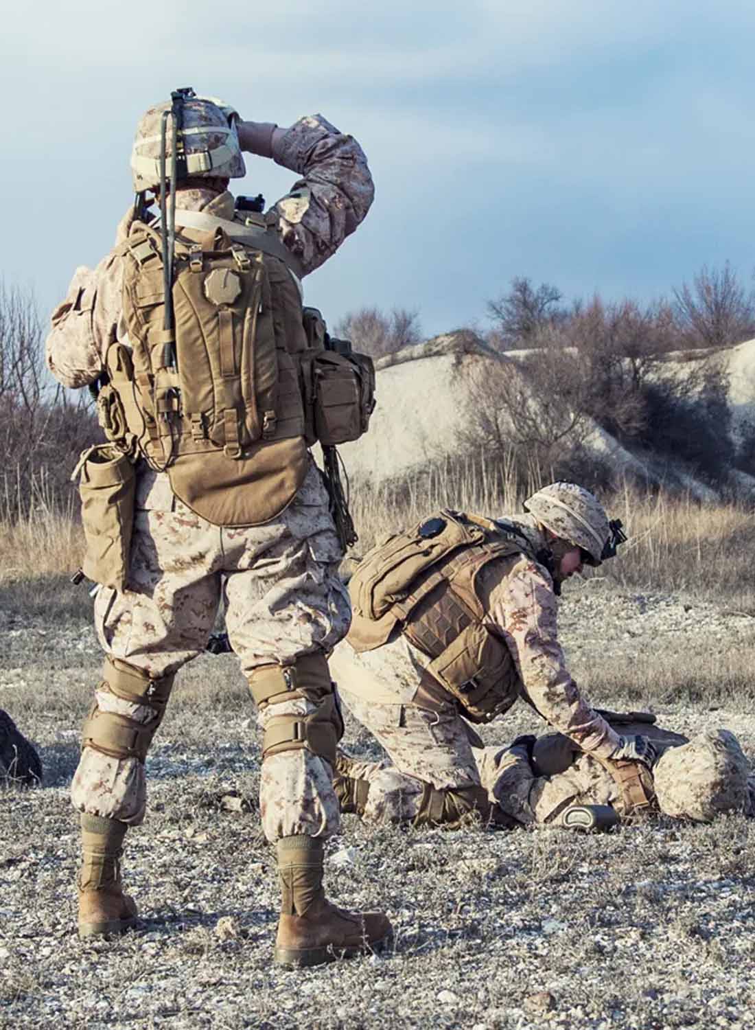 Photo shows the back of three soldiers outside, one is tending to the other lying on the group and the other is standing.