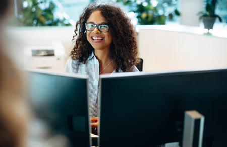 Photo shows a smiling woman with dark curly hair sitting behind two monitors.