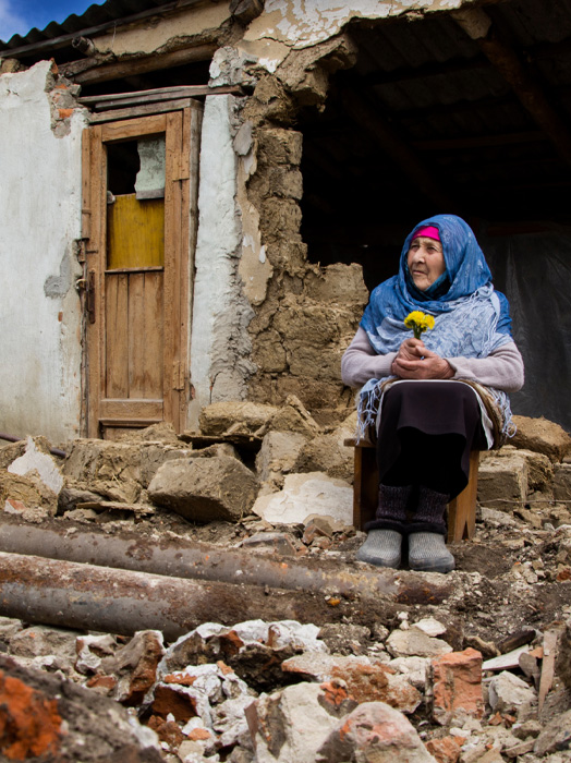 Photo shows an elderly woman holding flowers, sitting amongst a destroyed building.