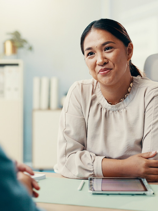 Photo shows a woman with dark hair in professional clothes behind a desk and speaking to someone out of shot.