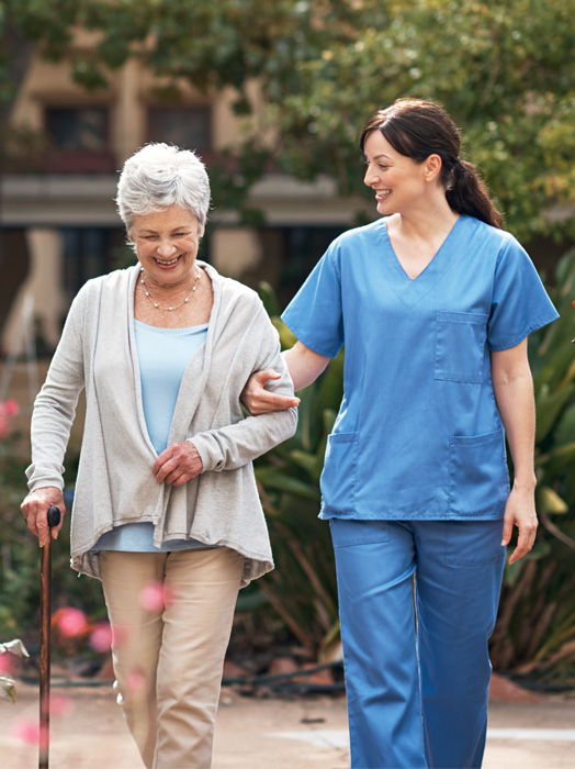 Photo shows a female clinician aiding an elderly woman with a walking stick as they take a stroll outside.