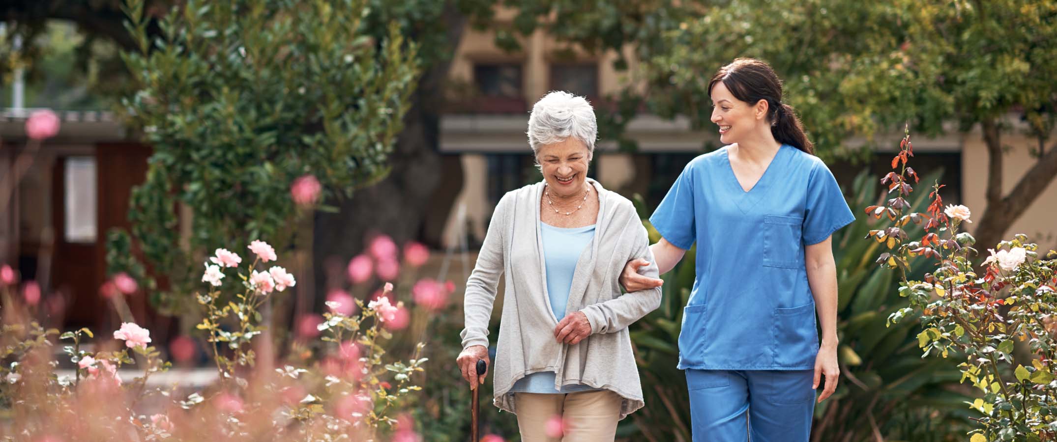 Photo shows a female clinician aiding an elderly woman with a walking stick as they take a stroll outside.