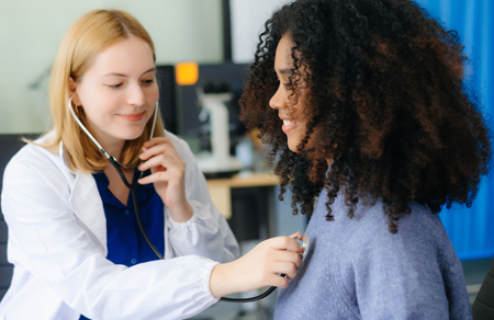 Photo displays a clinician holding the end of a stethoscope to the check of a female patient.