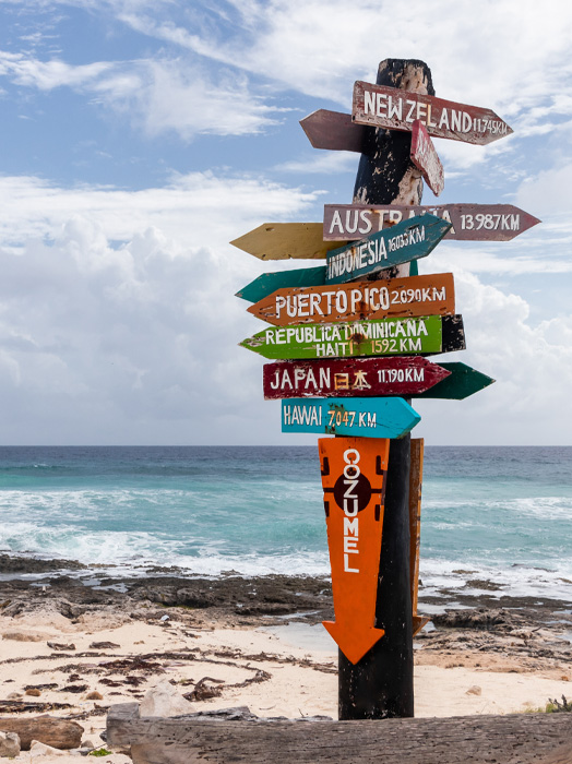Photo shows a wooden post at a beach with multiple directional signs to different places around the world.