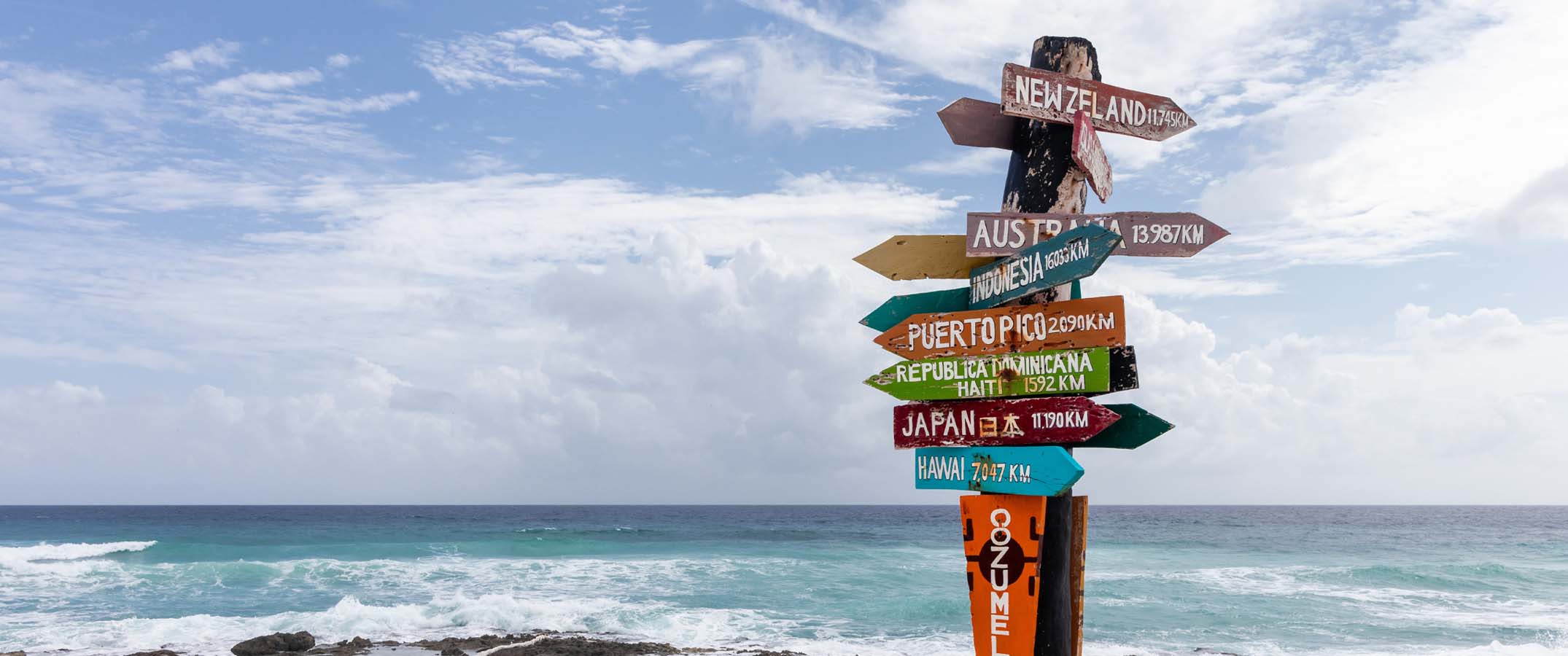 Photo shows a wooden post at a beach with multiple directional signs to different places around the world.