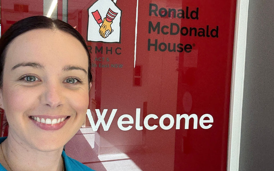 Photo of Casey smiling whilst standing in front of the welcome sign to Ronald McDonald House.