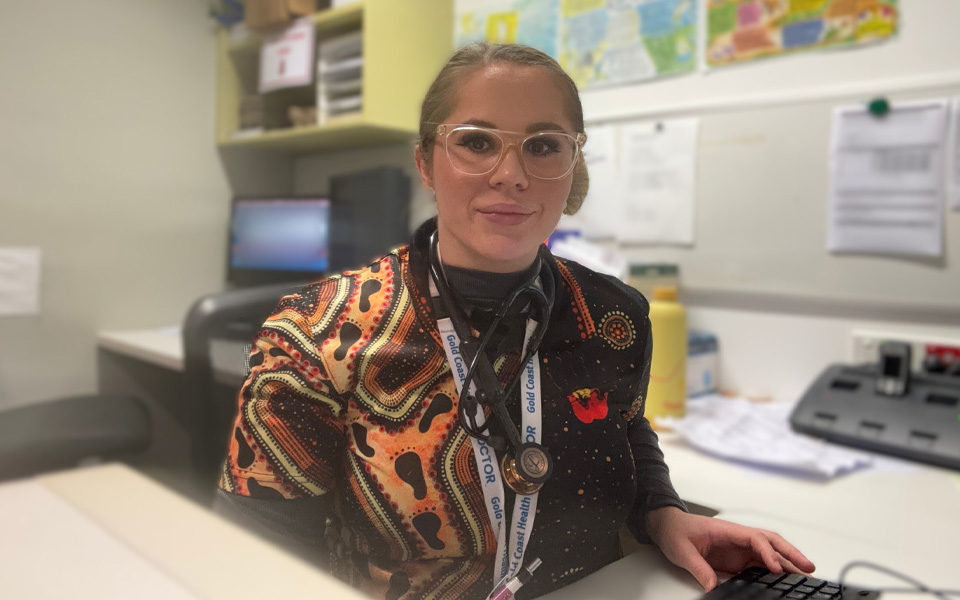 Photo of Dr Tahni Derbin sitting at her desk wearing an Indigenous artwork shirt.