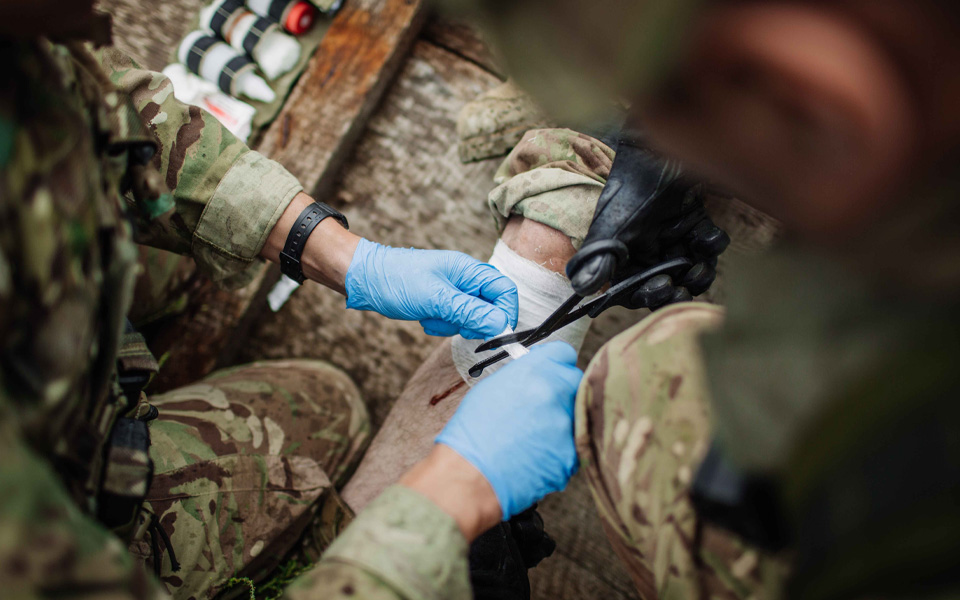 Image shows gloved hands cutting a bandage with people in defence uniforms.