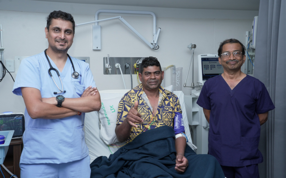 Two doctors in scrubs stand either side of a patient in a hospital bed