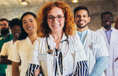 Stock image showing a clinician with red curly hair and glasses, smiling softly and standing infront of a group of people whose faces are slightly blurred.