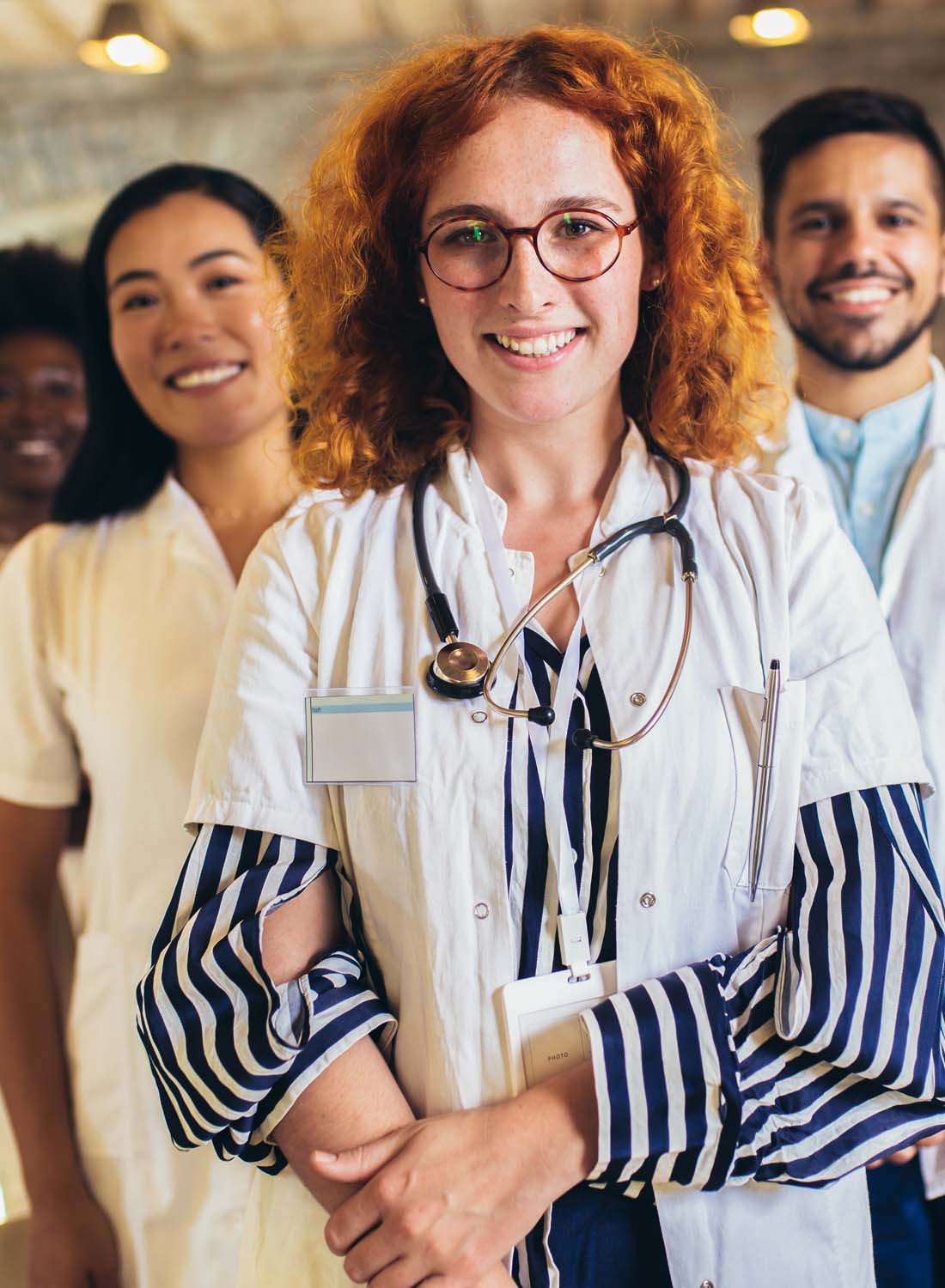 Stock image showing a clinician with red curly hair and glasses, smiling softly and standing infront of a group of people whose faces are slightly blurred.