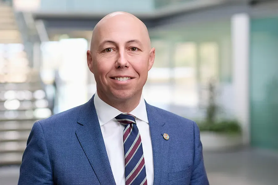 Al Martin, Chief Defence and National Security Officer at Aspen Medical, wearing a blue suit, white shirt and striped tie, with a lapel pin on his jacket, smiling in a professional office setting with a modern staircase and glass windows in the background