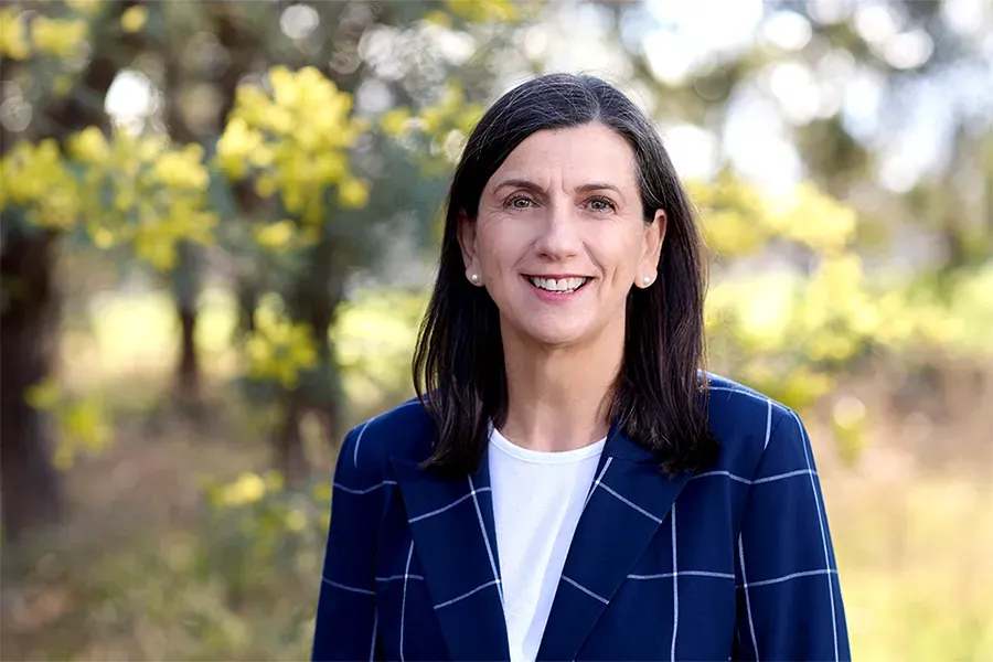 Mel Keys, Board Member of the Aspen Medical Foundation, wearing a navy blazer with white windowpane checks and a white top, smiling in a sunlit outdoor setting with yellow wattle flowers and native bushland in soft focus in the background.