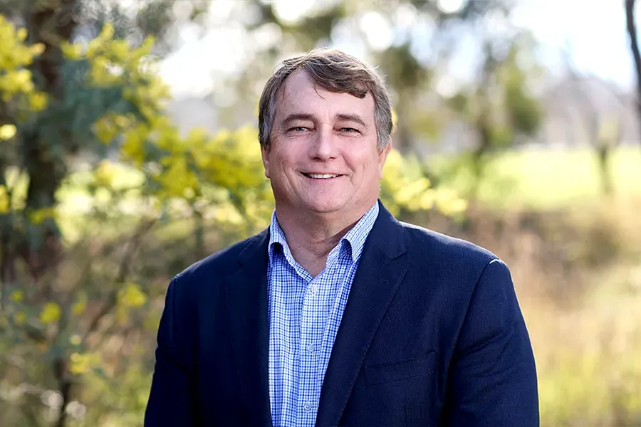 Craig Fitzgerald, Chief Executive Officer of the Aspen Medical Foundation, wearing a navy blazer and blue checkered shirt, smiling in a sunlit outdoor setting with yellow wattle flowers and native bushland softly blurred in the background.