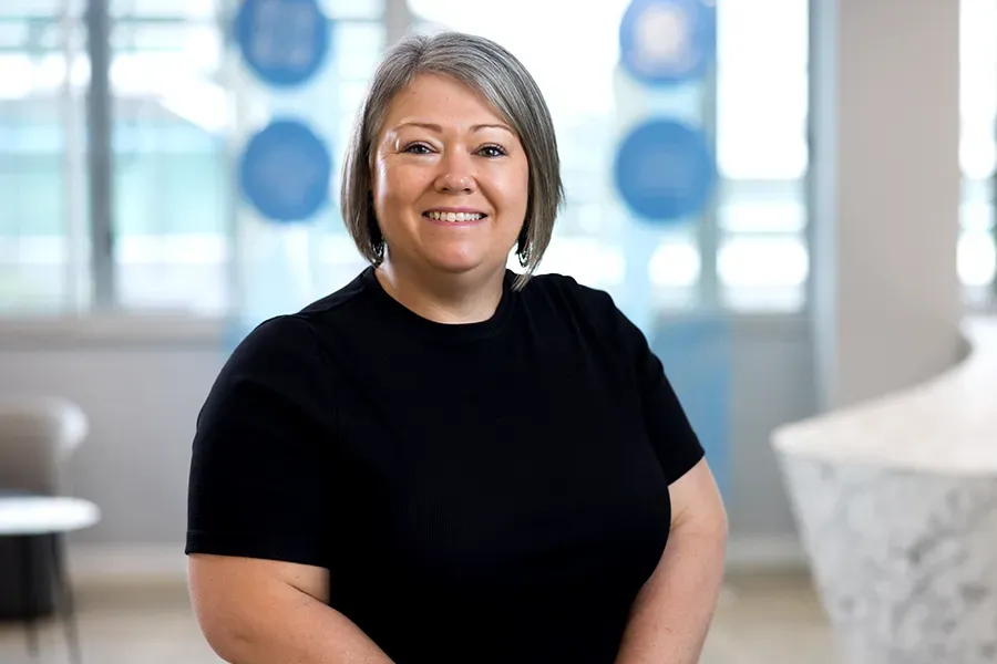 Dayle Mathews, Board Observer at the Aspen Medical Foundation, wearing a black top, smiling in a bright indoor setting with marble surfaces and blurred blue signage and windows in the background.