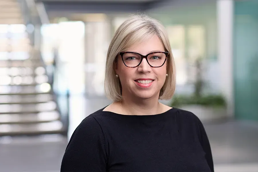 Emily Elliott, Chief People Officer at Aspen Medical, wearing a black top and black-framed glasses, with short blonde hair, smiling in a professional office setting with a modern staircase and glass windows in the background.