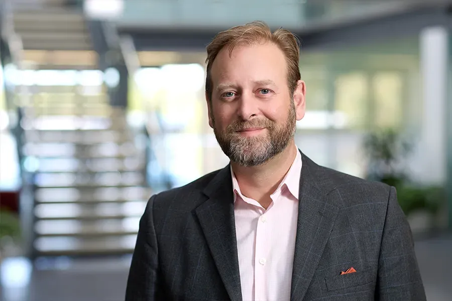 Ethan Bond, President of Aspen Medical USA, wearing a dark grey checked suit jacket and light pink shirt, standing in a professional office setting with a modern staircase and glass windows in the background.