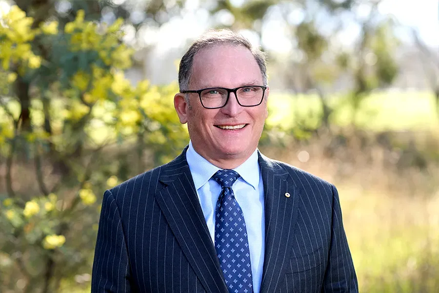 Glenn Keys AO, Board Member of the Aspen Medical Foundation, wearing a navy pinstripe suit, light blue shirt and navy patterned tie, smiling in a sunlit outdoor setting with yellow wattle flowers and soft-focus native bushland in the background.