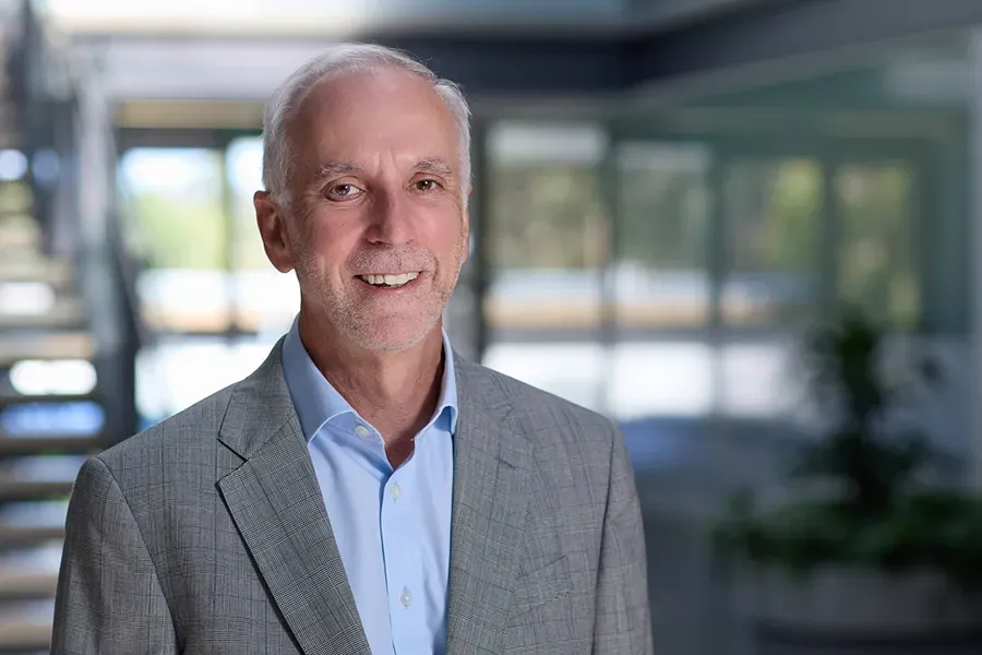 John Stevenson, Chief Financial Officer of Aspen Medical, wearing a light grey checkered blazer over a light blue shirt, smiling in a professional office setting with a modern staircase and glass windows in the background.