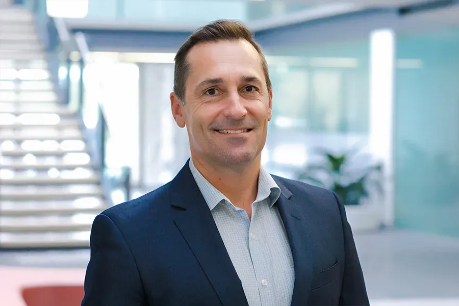 Matt Hughes, Chief Strategy Officer of Aspen Medical, wearing a navy blazer over a light blue checkered shirt, smiling in a professional office setting with a modern staircase and glass windows in the background.
