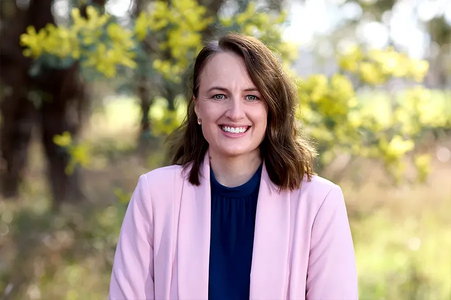 Melissa Broers, Social Impact Manager at the Aspen Medical Foundation, wearing a soft pink blazer and navy blouse, smiling in a sunlit outdoor setting with yellow wattle flowers and softly focused native bushland in the background.
