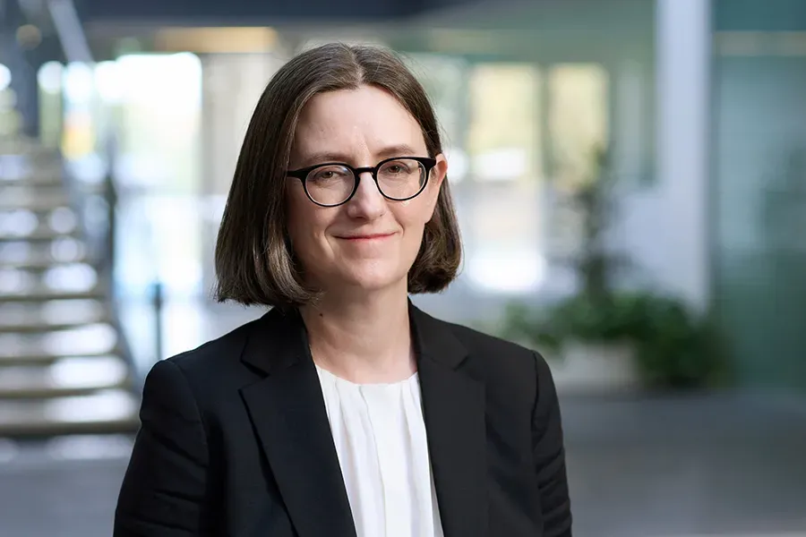 Michelle Widmaier, General Counsel at Aspen Medical, wearing a black blazer over a white blouse, with shoulder-length brown hair and black-framed glasses, smiling in a professional office setting with a modern staircase and glass windows in the background.