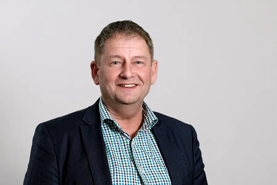 Paul Bird, Board Member of the Aspen Medical Foundation, wearing a navy textured blazer and a green, blue and white checkered shirt, smiling in a professional indoor setting with a plain light-grey background.